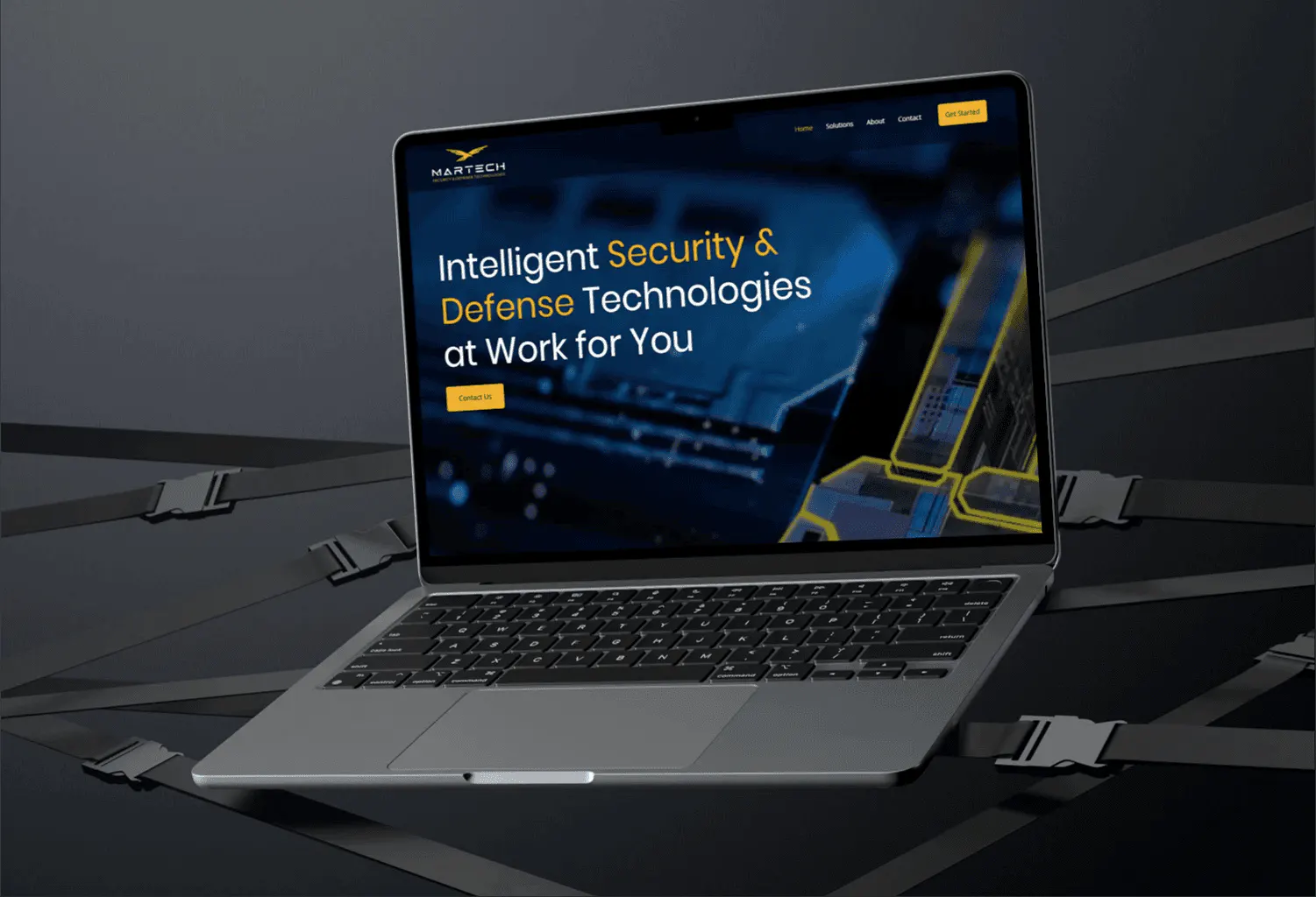 A laptop resting on crisscrossed black straps with buckles, on a dark surface, showing the MARTECH home page headline 'Intelligent Security & Defense Technologies at Work for You'