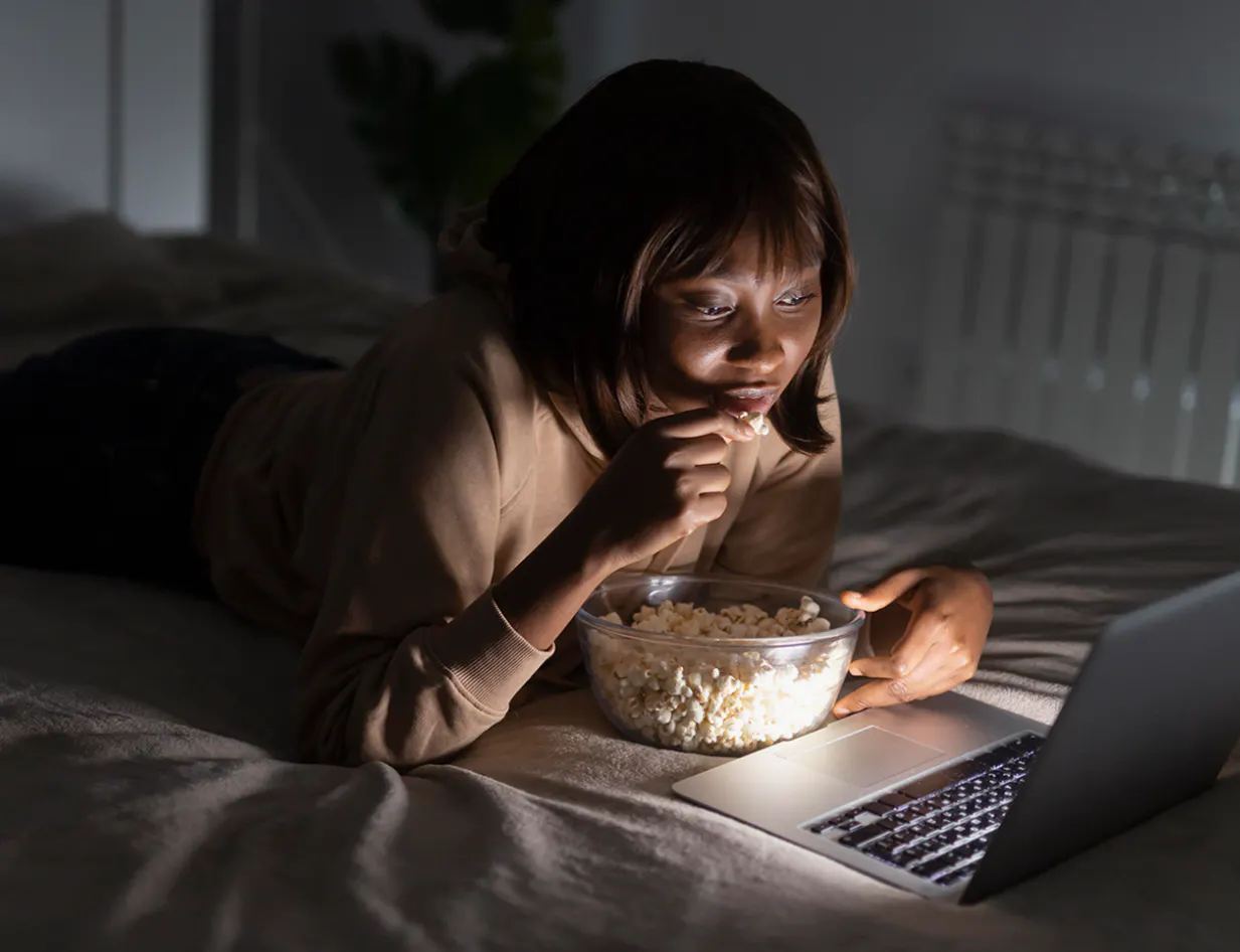 Photograph of a woman lying on a bed at night, eating popcorn and watching a laptop