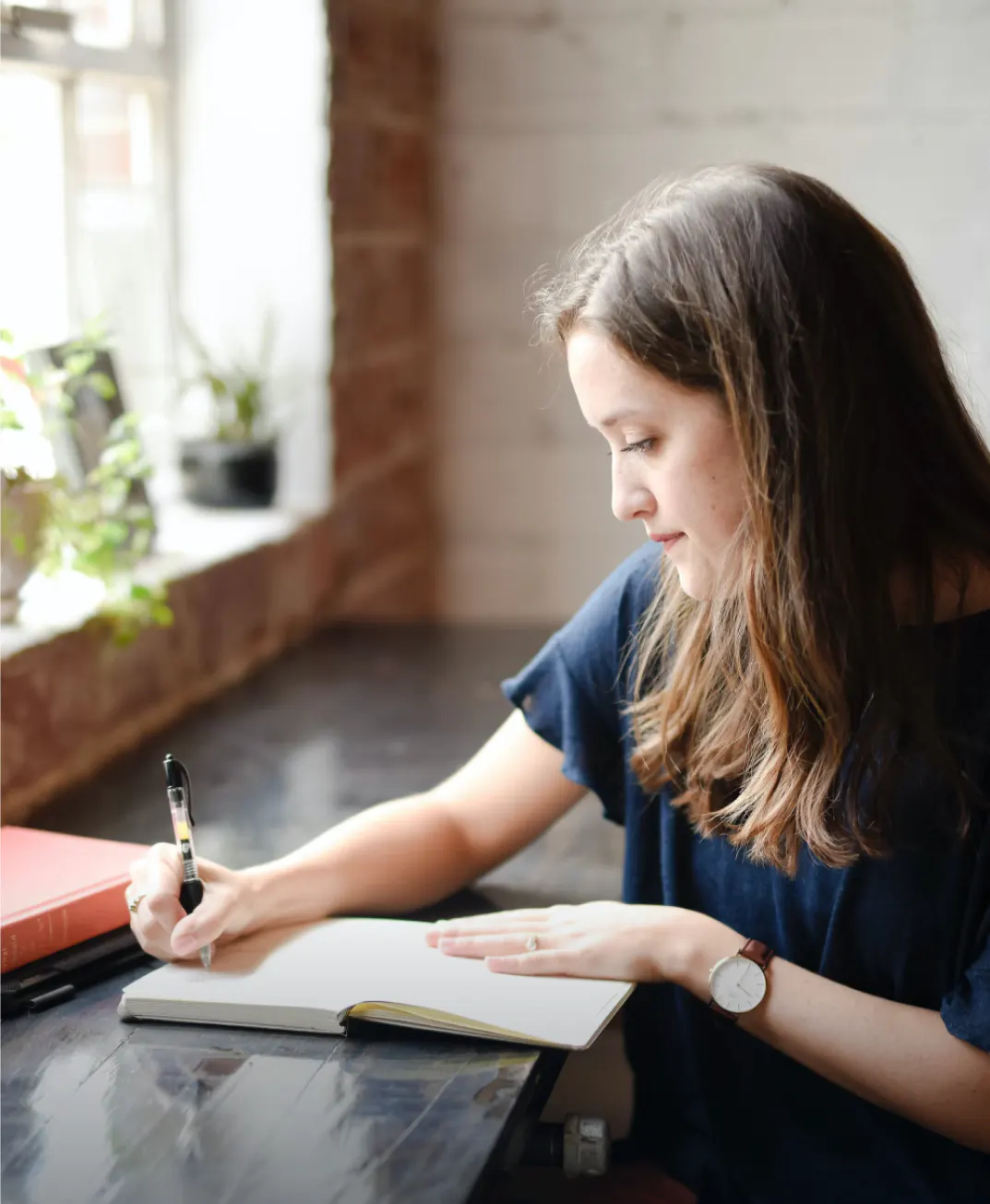 Camille’s photo: a young woman writing in a notebook at a dark wooden table by a window, with plants on the sill